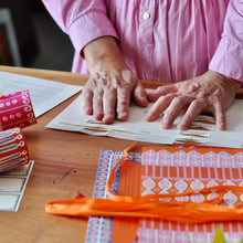 A close-up of a person's hands arranging patterned paper strips on a wooden table, with sheets of patterned paper and orange ribbon in the foreground.