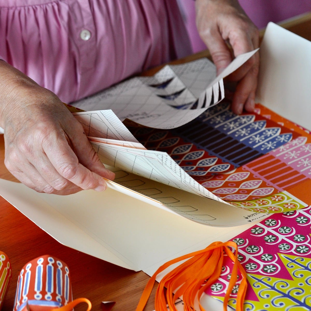 A close-up of hands folding patterned paper sheets on a wooden table, with colourful printed paper stacks and orange ribbon visible in the scene.