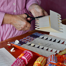 A person is cutting out a white paper template with zigzag perforations, working over a wooden table with colourful patterned paper pieces to make Christmas crackers.