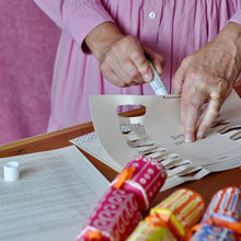A person is using a glue stick form the crackers on a wooden table, with printed papers and coloruful finished crackers in the foreground.