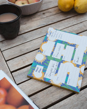 A colorful printed meal plan notepad with illustrated sunflower background, placed on a wooden table with a mug, fruit, and a cookbook.