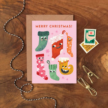 Christmas card with colourful happy stockings on a pink background, placed on a wooden surface with decorative items.