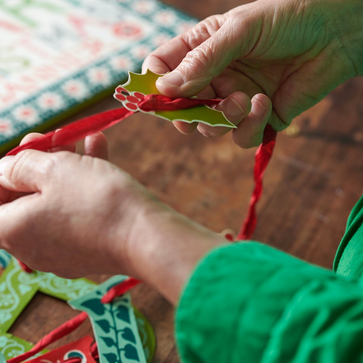 Close-up of hands threading a holly leaf with berries onto Merry Christmas banner by Cambridge Imprint.