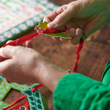 Close-up of hands threading a holly leaf with berries onto Merry Christmas banner by Cambridge Imprint.