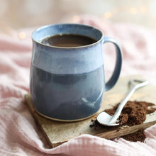 Blue handmade ceramic mug filled with coffee on a wooden coaster with a spoon and grounds, on a soft pink fabric background.