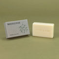 A bar of Mistletoe natural soap bar in an off-white cream color, shown next to its textured gray packaging box with a green mistletoe illustration, set against a solid olive green background.