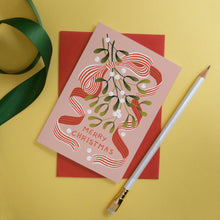 A pink Christmas greeting card featuring mistletoe and striped red and white ribbon with a red envelope, styled on a yellow background with a green ribbon and a white pencil.