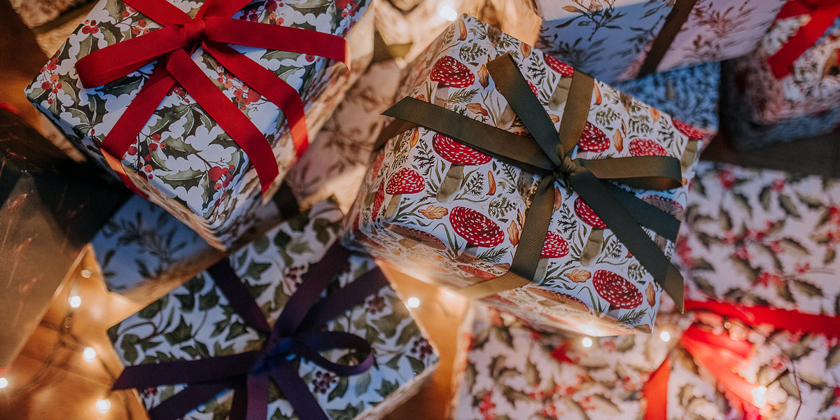 Several wrapped Christmas gift boxes with patterned paper and large ribbon bows, including red, dark green, and purple ribbons, arranged on a wooden surface with warm string lights.