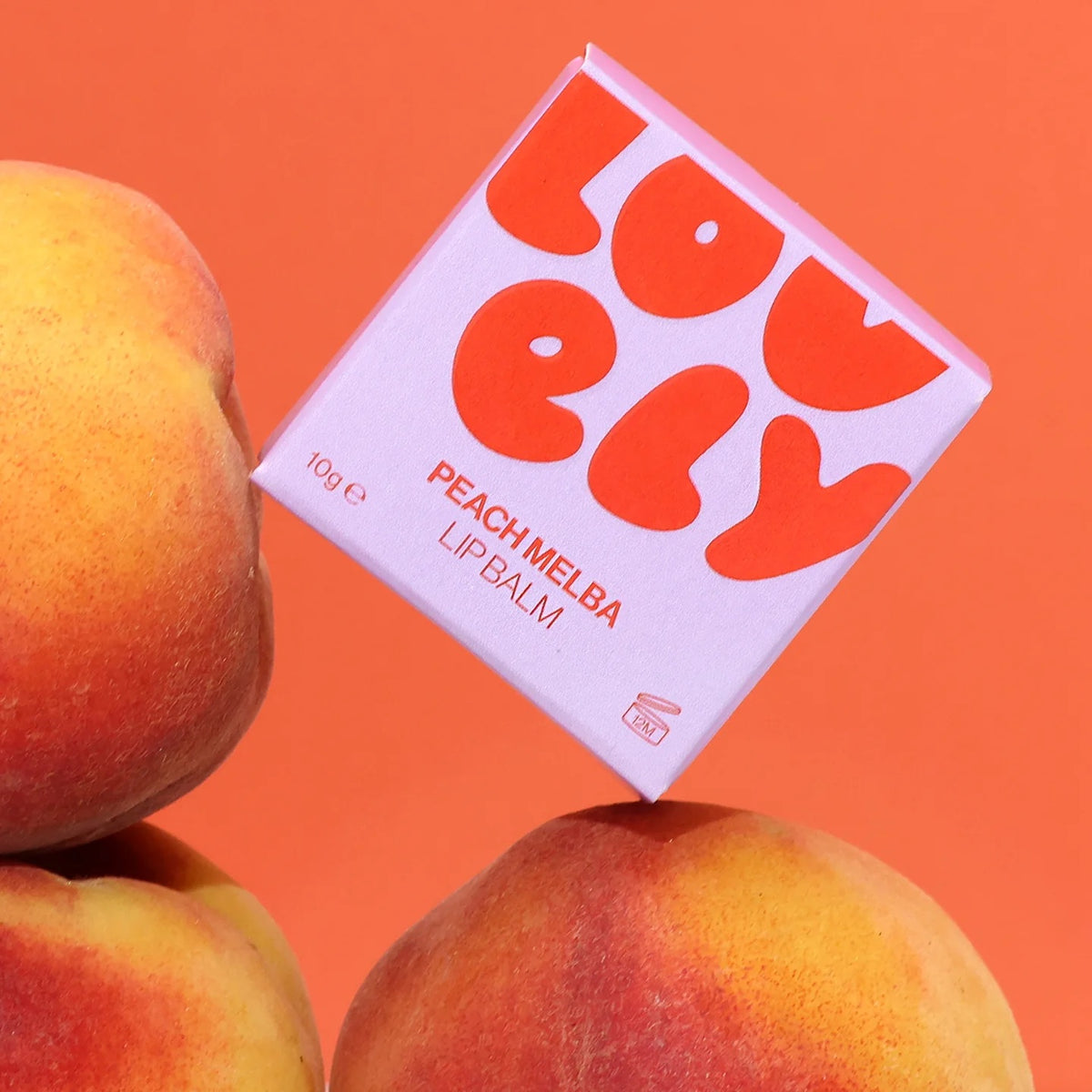 A square box of Lovely Skincare Peach Melba lip balm is shown tilted on top of peaches against a solid orange background.