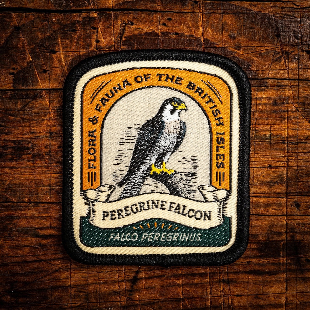 A rectangular embroidered patch with rounded corners featuring a peregrine falcon illustration and the text 'Flora & Fauna of the British Isles' and 'Peregrine Falcon', placed on a rustic wooden surface.
