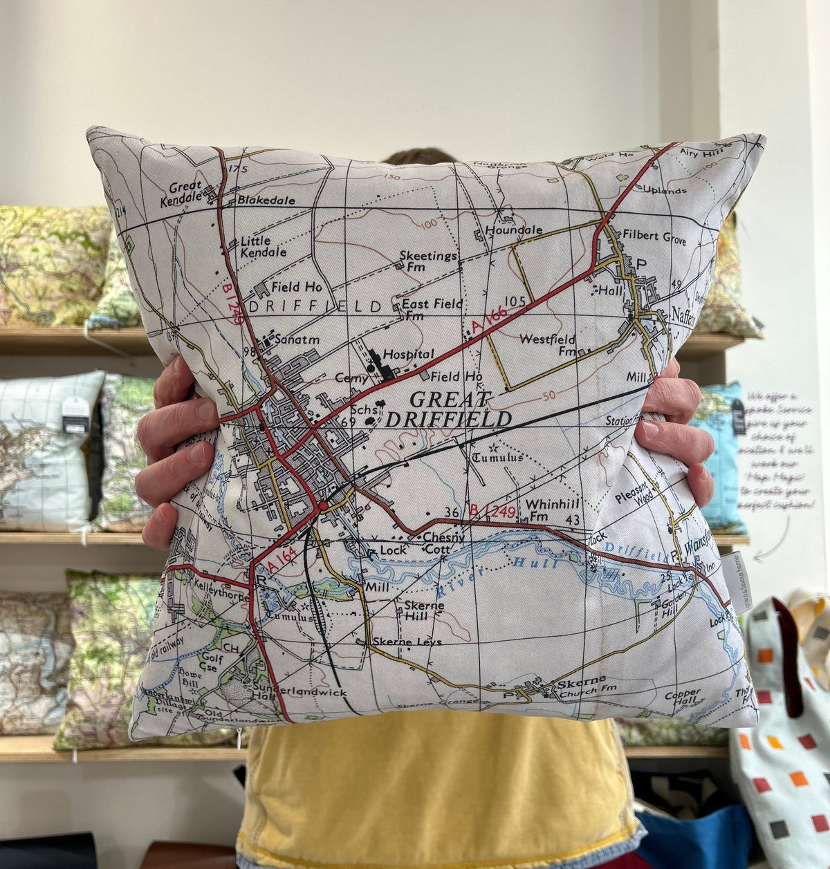 A person holds up a square throw pillow printed with a vintage-style map of Great Driefield, showing their hands and torso. The background shows a shop interior with shelves of other pillows.