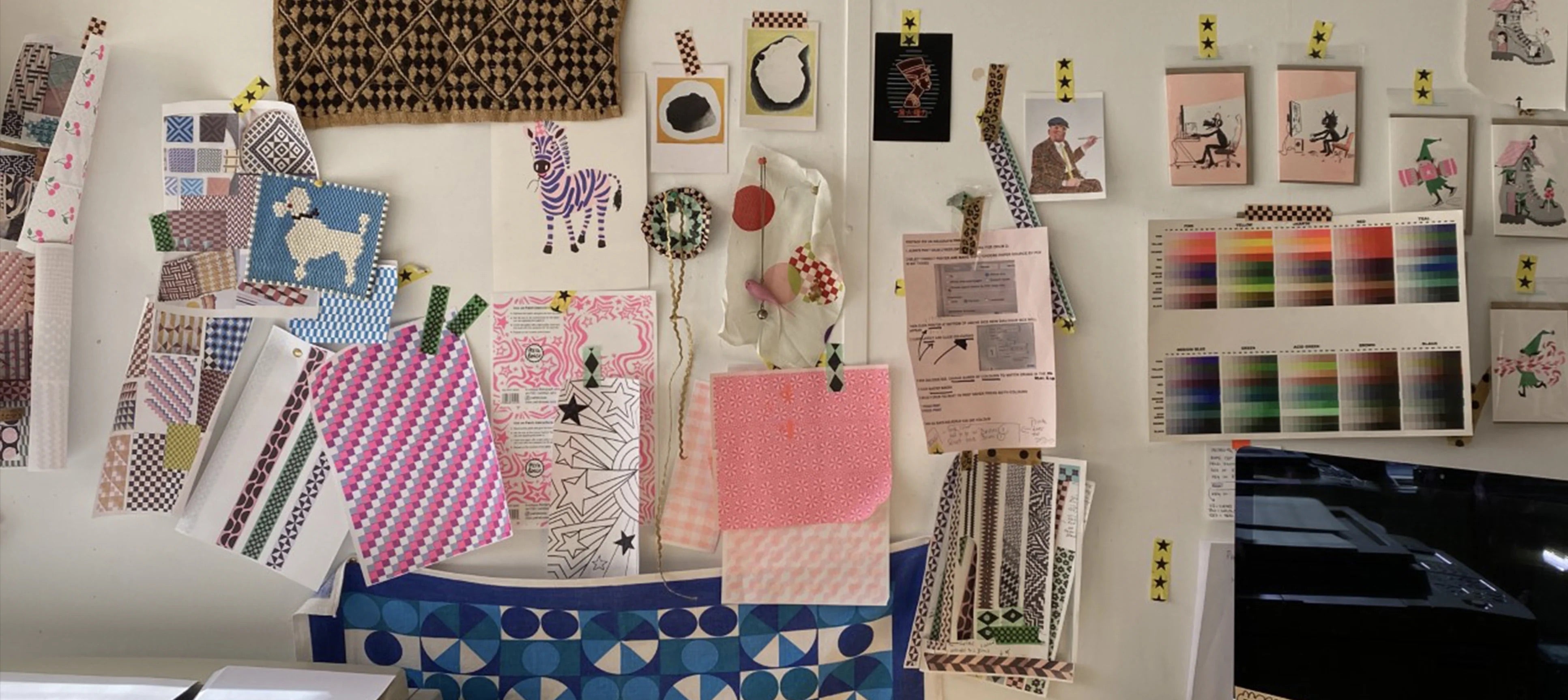 A display of various fabric swatches, patterned paper samples, small framed art prints, and handmade textile pieces pinned to a white wall in an art studio or craft room.