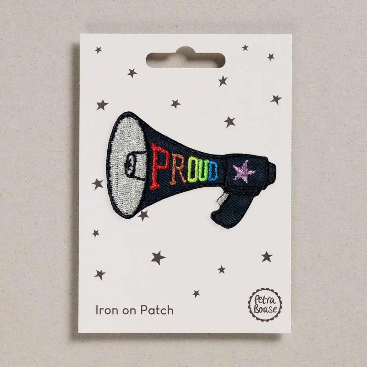 An embroidered iron-on patch shaped like a megaphone with the word 'PROUD' in rainbow colors and a pink star, displayed on a white backing card with star graphics. The backing card is labeled 'Iron on Patch' and shows the brand 'Petra Boase'.