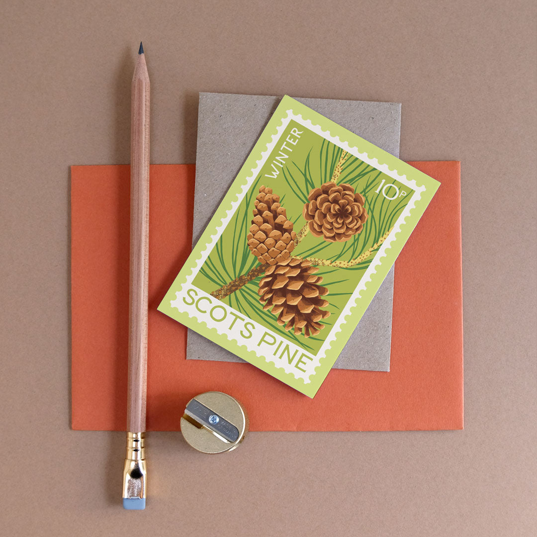 A postage stamp-style card with pinecone artwork, arranged on orange and gray paper with a wooden pencil and a small sharpener on a taupe background.