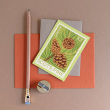 A postage stamp-style card with pinecone artwork, arranged on orange and gray paper with a wooden pencil and a small sharpener on a taupe background.