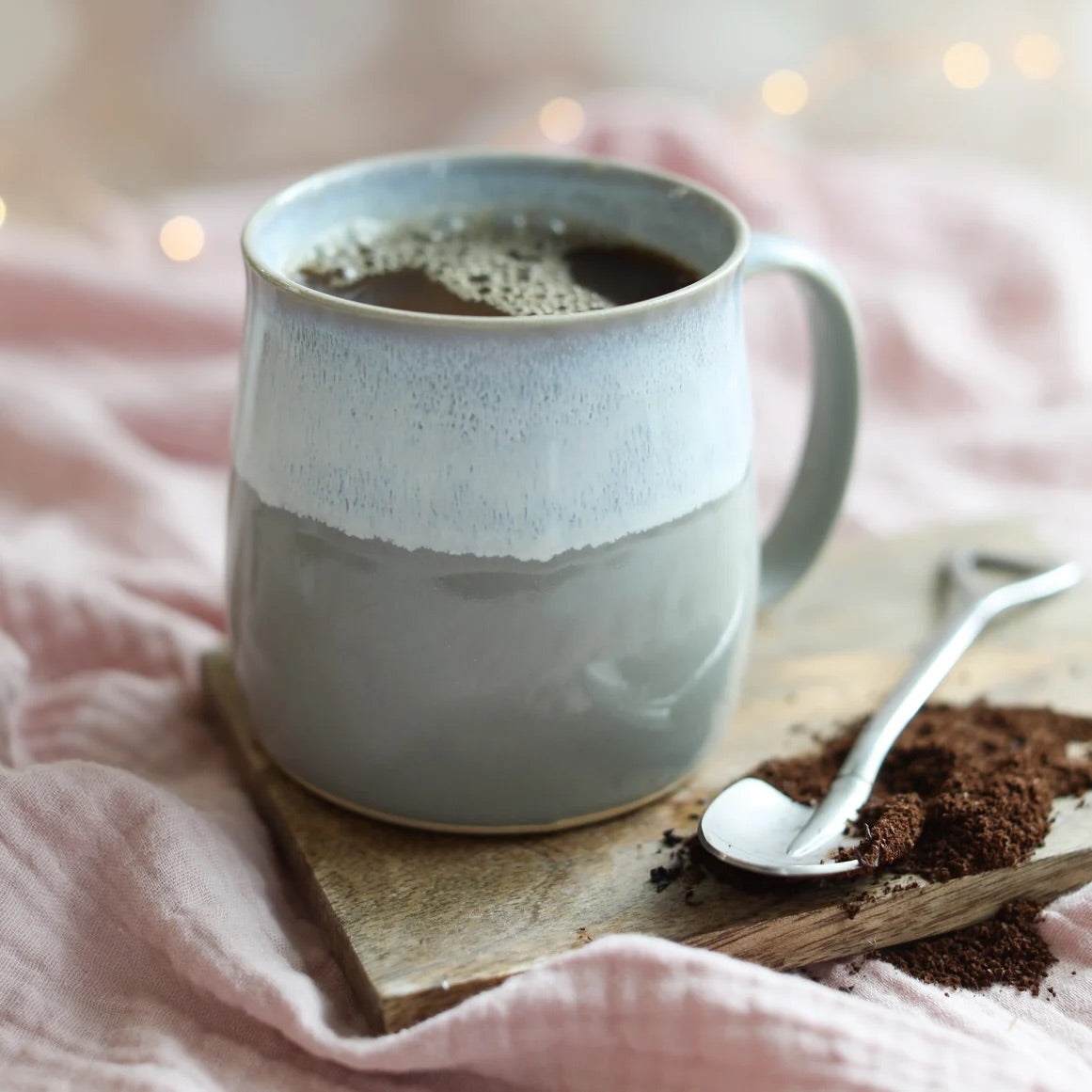 Ceramic mug with coffee on a wooden coaster with a spoon and grounds, against a soft pink background.