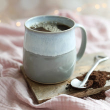 Ceramic mug with coffee on a wooden coaster with a spoon and grounds, against a soft pink background.