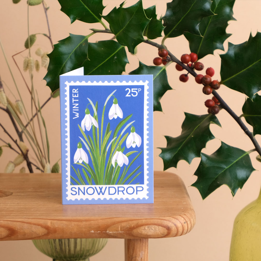 Mini card featuring snowdrop vintage stamp style design in blue, shown on wooden bench with holly in the background.