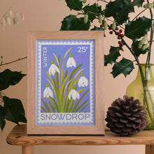 A framed art print featuring a vintage stamp style illustration of white snowdrops on a lavender background, styled on a wooden table with a pinecone and a glass vase of holly leaves.