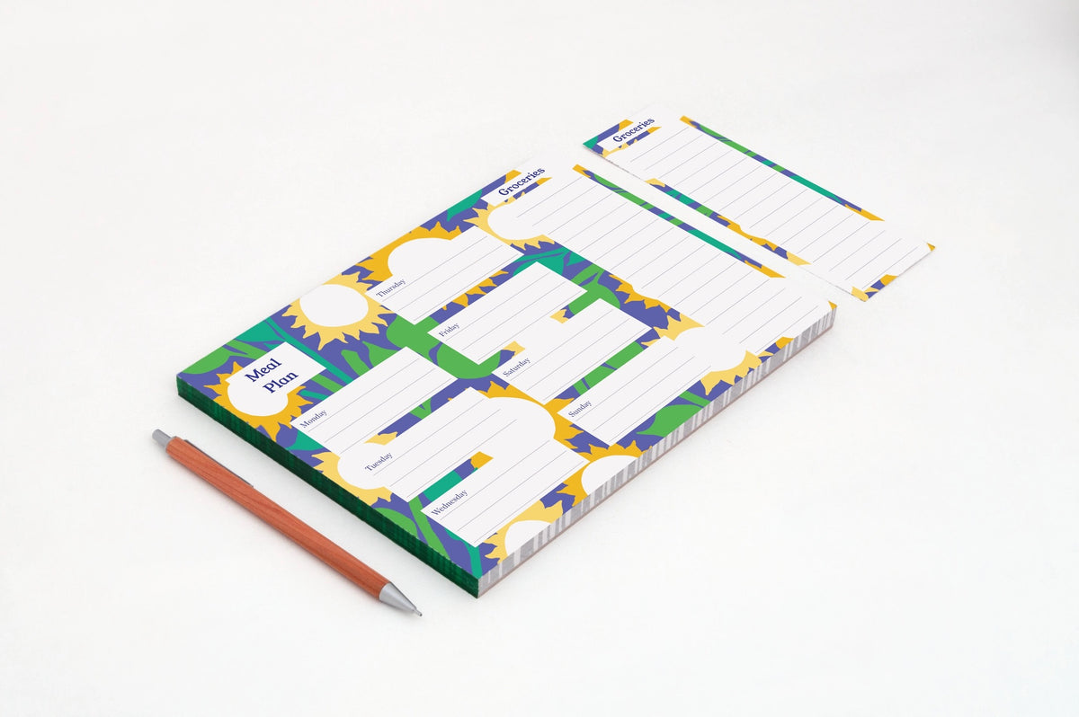A meal planning pad with a colorful sunflower pattern background, with a brown pencil placed beside it on a clean white background.