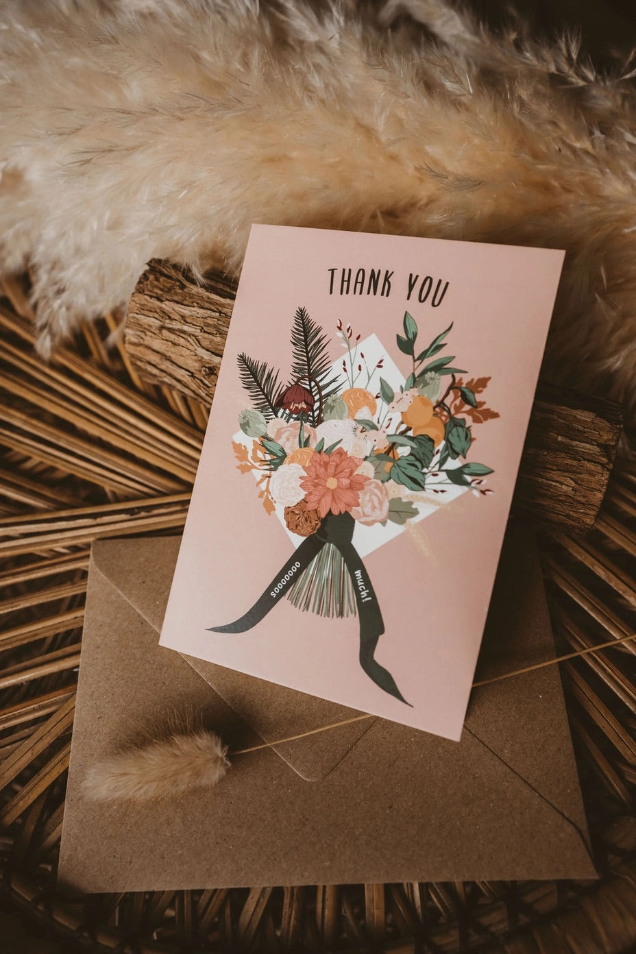A pink thank you card with a floral bouquet illustration and the words 'THANK YOU' at the top and 'sooooooo much!' on the ribbon tails, placed on a brown envelope.