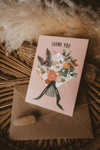 A pink thank you card with a floral bouquet illustration and the words 'THANK YOU' at the top and 'sooooooo much!' on the ribbon tails, placed on a brown envelope.