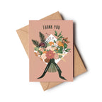 A thank you card with a dusty pink background and an illustrated bouquet of flowers tied with a dark green ribbon, shown with a brown kraft envelope on a white background.