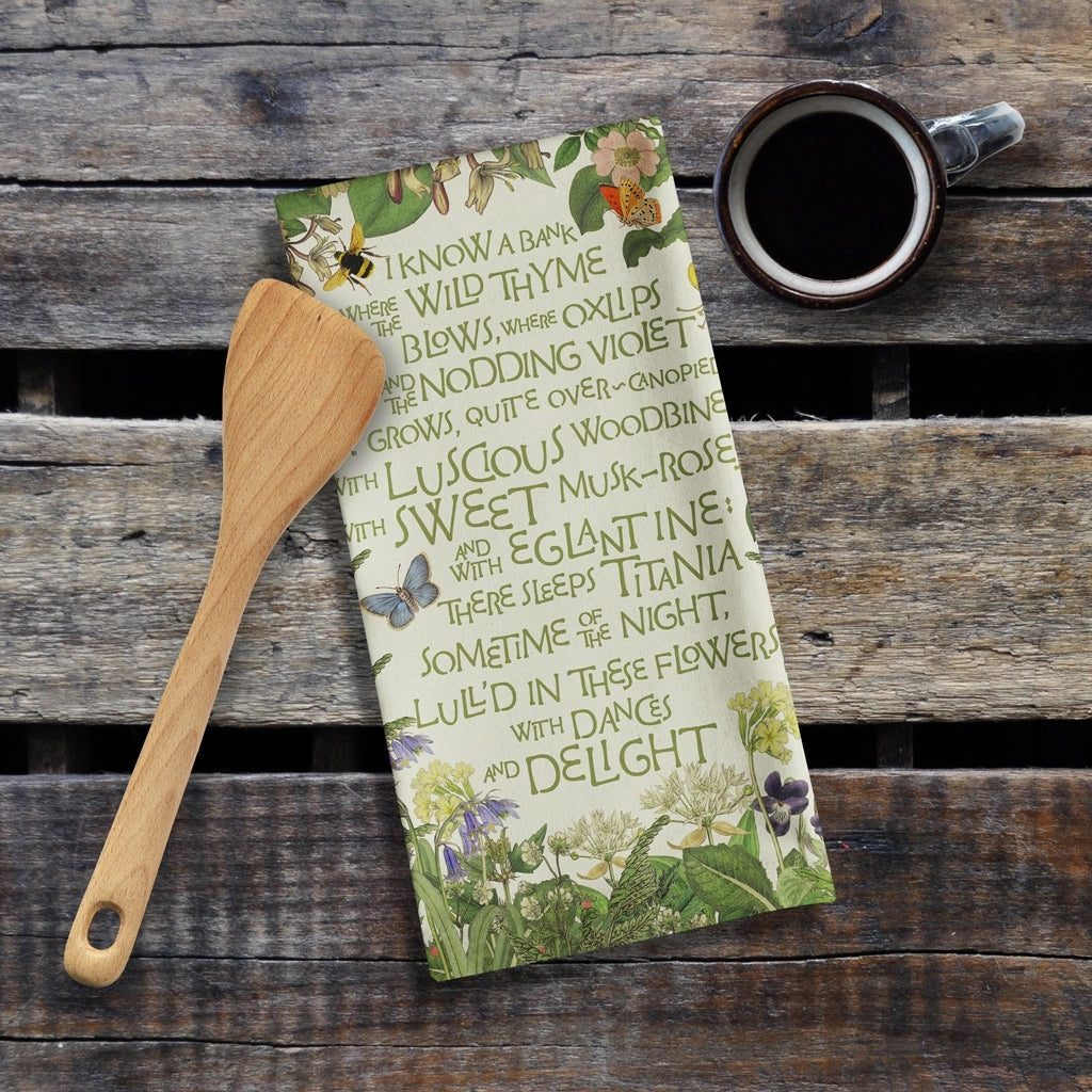 Tea towel with floral and text design on a wooden surface with a wooden spatula and cup of coffee.