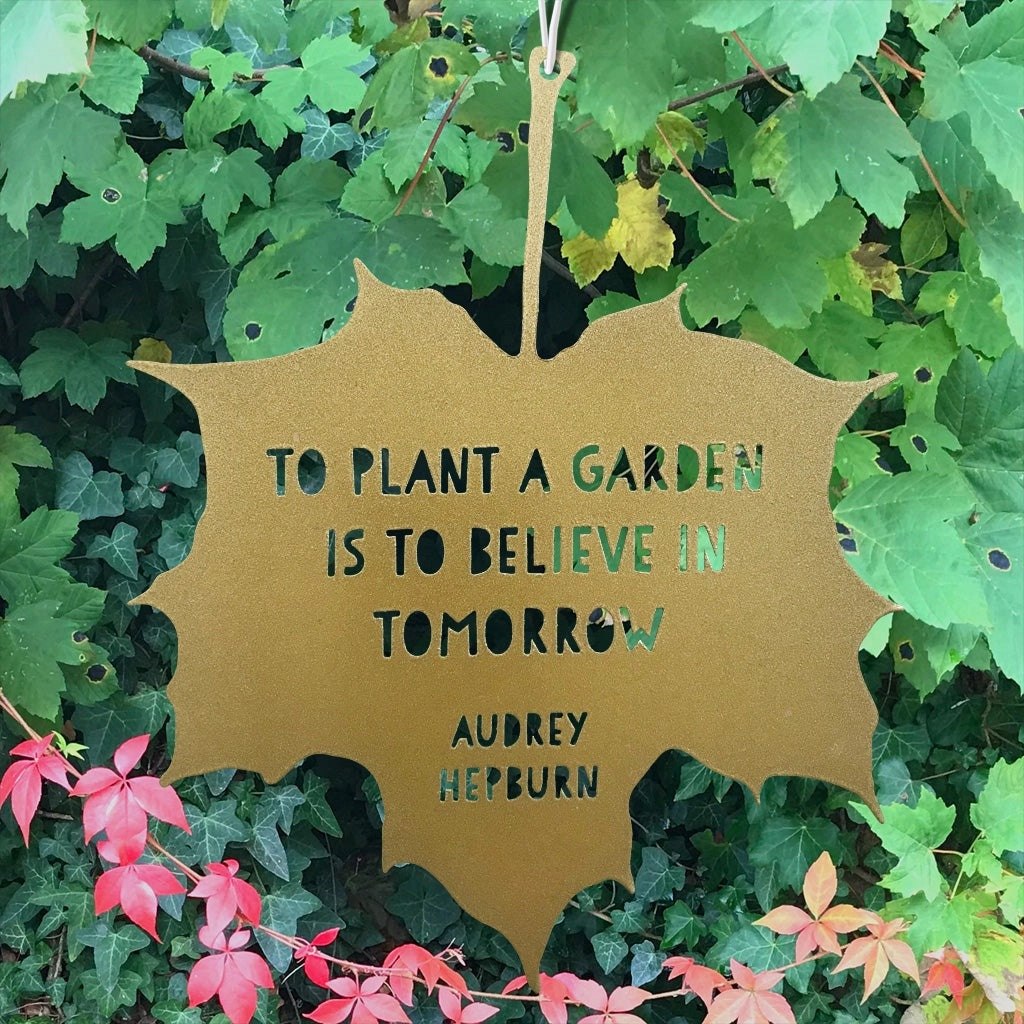 Gold leaf-shaped sign with a quote by Audrey Hepburn hanging among green leaves.