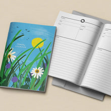 A travel logbook shown closed and open on a light beige surface. The closed cover features a colorful illustration of tall green grass, white daisies, purple flowers, a yellow sun, and a blue sky with small birds.