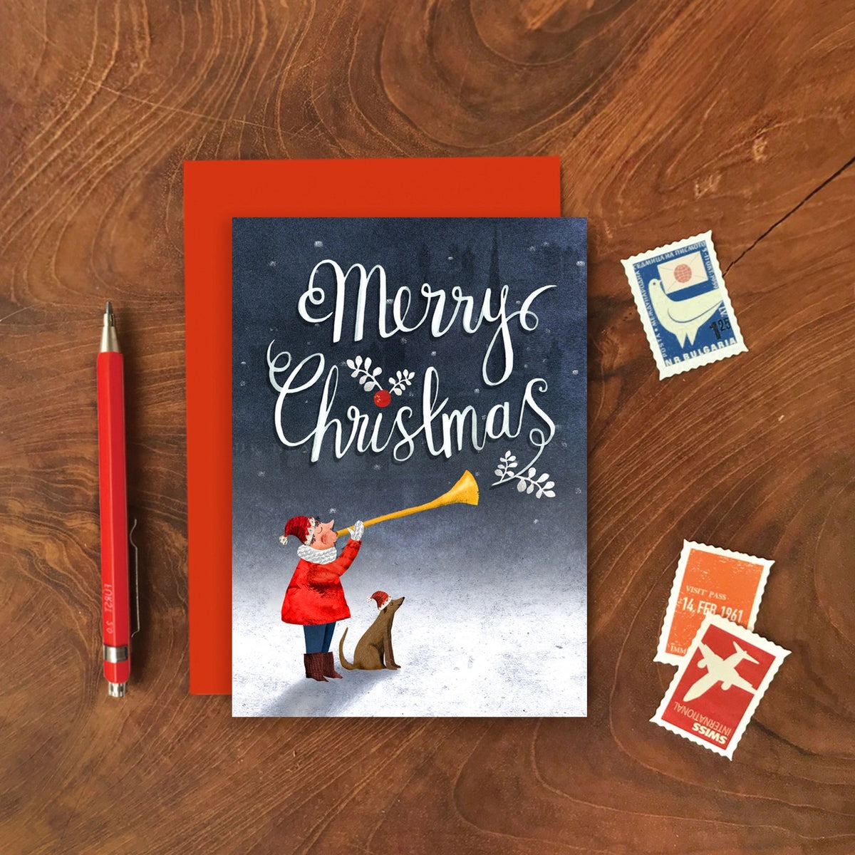 Christmas card with trumpeter and dog illustration on a wooden surface with stamps and a pen.