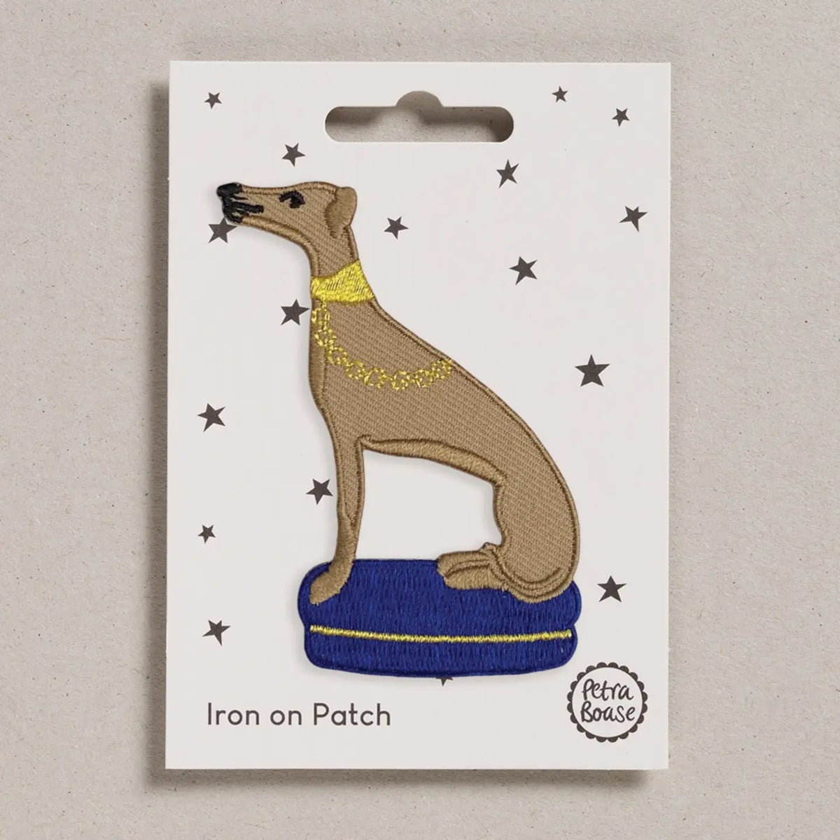 An embroidered iron-on patch of a whippet dog sitting on a purple/blue cushion with a yellow collar, displayed on its white backing card with star details.