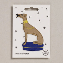 An embroidered iron-on patch of a whippet dog sitting on a purple/blue cushion with a yellow collar, displayed on its white backing card with star details.