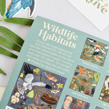 A calendar back page titled 'Wildlife Habitats' laid on a white surface with decorative green leaves and a partial card around it. The calendar has a muted blue-green background and contains illustrated wildlife scenes, including birds, fish, insects, and sea life, with text describing an annual calendar focused on UK habitat.