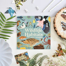 A square 2027 Wildlife Habitats calendar lies on a white surface surrounded by decorative natural props, including leaves, a wood slice, paper clips, and butterfly and bird stickers. The calendar cover features a light blue background with many illustrated animals and habitats, and the title 'Wildlife Habitats 2027 Calendar' is printed prominently.