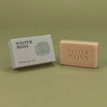 A bar of natural soap bar next to its textured gray packaging box, both embossed with the brand name 'WINTER MOSS', set against a solid olive green background.