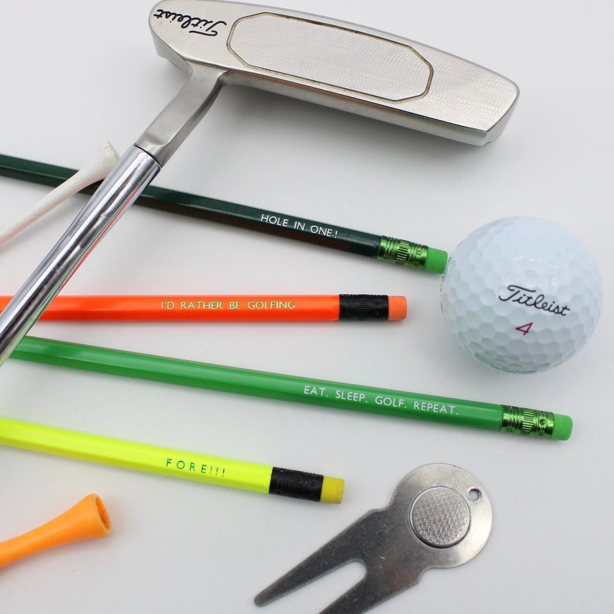 Four pencils with golfing terms are displayed with a set of golf accessories arranged on a white background, including a silver putter, a white Titleist golf ball,.