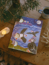 Advent calendar featuring a whimsical illustration of puffins, and houses by the sea, styled on a rustic wooden table with a candle, mug, and greenery.
