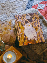 Advent calendar featuring a white owl perched among bare trees under a full moon, styled on a knitted blanket with a quilted textile and a lit candle nearby.