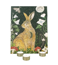 Advent calendar featuring a detailed illustration of a brown hare in a forest at night, surrounded by white flowers, green leaves, red-and-white mushrooms, a butterfly, and a full moon. Three small tealight candles are placed in front on a white background.